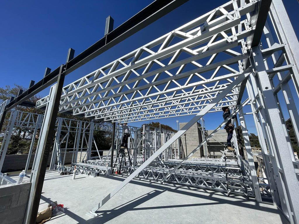Steel wall frames and roof trusses being assembled on a building site under a clear sky.