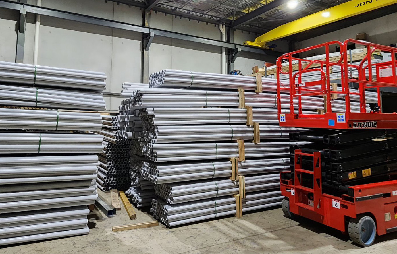 Bundles of light-coloured pipes stacked on timber gluts in a warehouse, beside a red scissor lift.